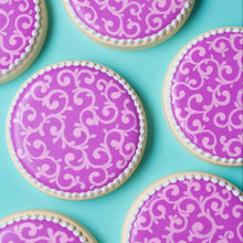 round cookies decorated with bright purple royal icing and a light purple filigree design. There is a white bead border on the edges of the cookies. The cookies are on a teal background.